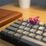 Figura de axolote color rosa estilo pixel art posada sobre las teclas de un teclado mecánico gris en un escritorio gamer.