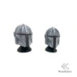 Vista en perspectiva de dos réplicas de cascos de Star Wars sobre soportes negros, destacando el acabado metalizado.