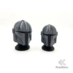 Comparativa de dos tamaños de cascos Mandalorianos color plata sobre bases negras, fondo blanco limpio.