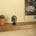 Casco del Mandaloriano decorando una repisa de madera rústica con un póster vintage del Halcón Milenario de Star Wars al fondo.
