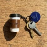 Llavero con forma de vaso de café realista marca Pracktico sobre superficie de madera con llaves.